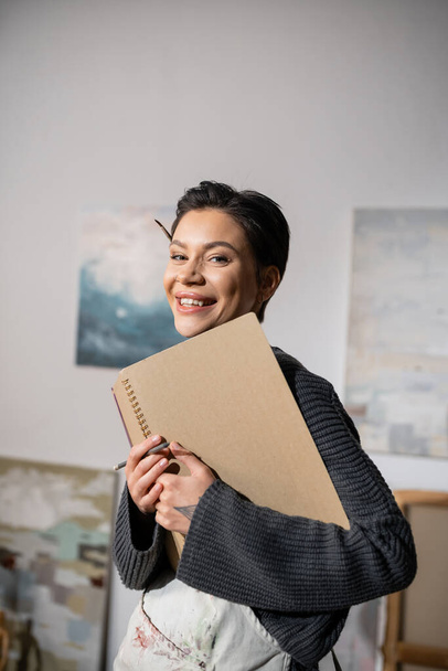 Portrait of cheerful artist in apron looking at camera while holding sketchbook  - Foto, Imagen