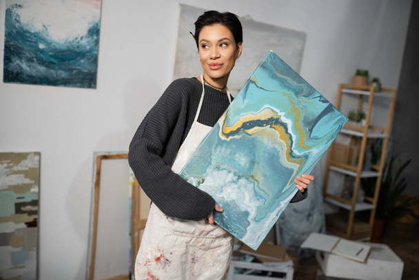 Pleased artist in apron looking away while holding painting in workshop  - Фото, зображення
