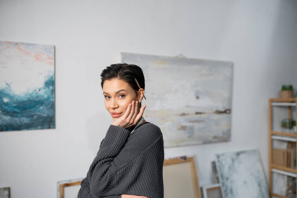 Smiling brunette artist with paintbrush in hair looking at camera in studio  - Valokuva, kuva