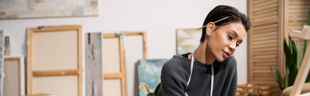 Lyhytkarvainen taiteilija siveltimellä katselee maalaustelinettä studiossa, banneri  - Valokuva, kuva