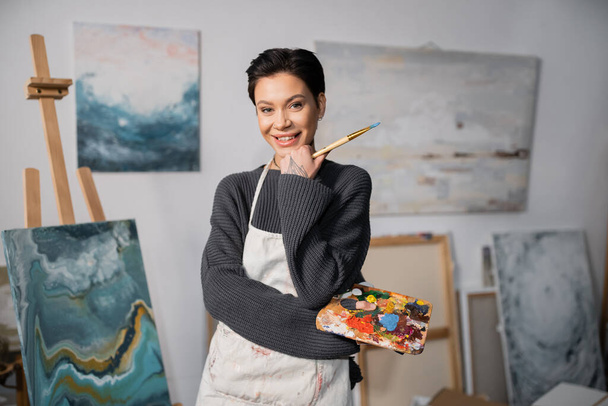 Cheerful tattooed artist holding paintbrush and palette in studio  - Zdjęcie, obraz
