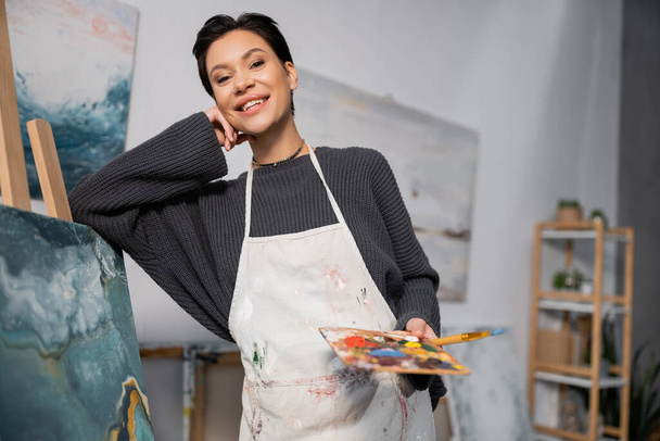 Smiling artist in dirty apron holding paintbrush and palette near painting in studio  - Fotoğraf, Görsel