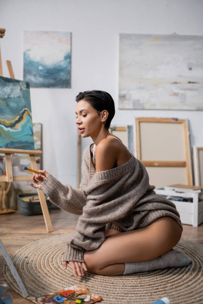 Sexy artist holding paintbrush near palette on floor in studio  - Fotó, kép