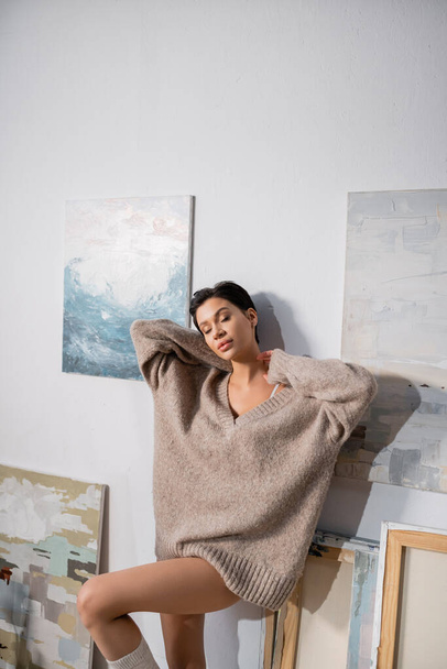 Sensual artist in sweater standing near drawings on wall  - Foto, immagini