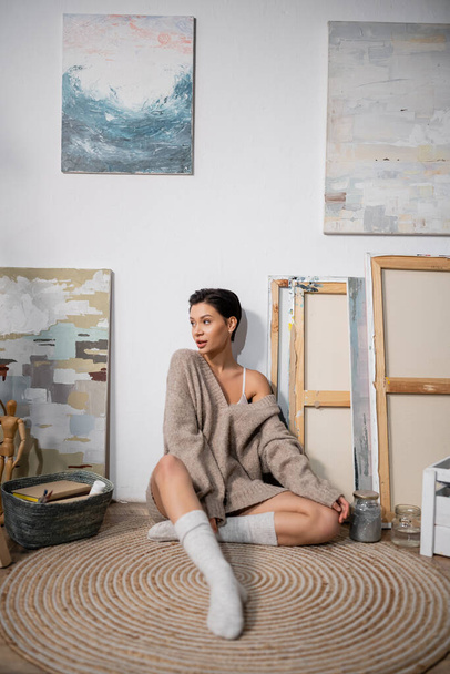 Short haired artist in sweater sitting near paintings on floor in workshop  - Foto, immagini