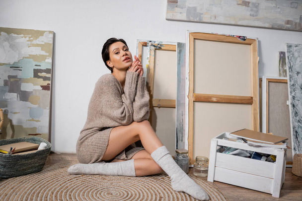 Sexy brunette artist sitting near paintings in workshop  - Foto, Imagen