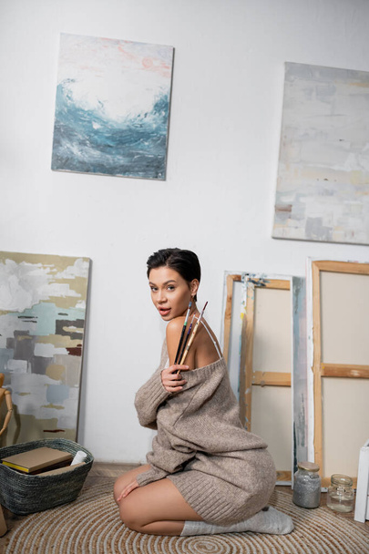 Seductive brunette artist holding paintbrushes while sitting on floor in workshop  - Fotografie, Obrázek