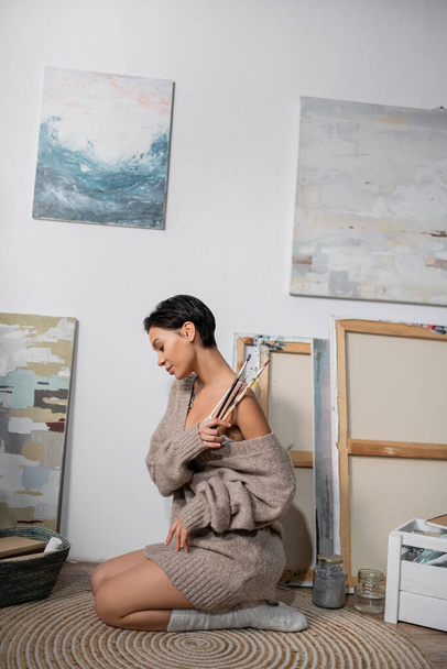 Side view of brunette artist in sweater and bra holding paintbrushes near drawings in workshop  - Photo, Image