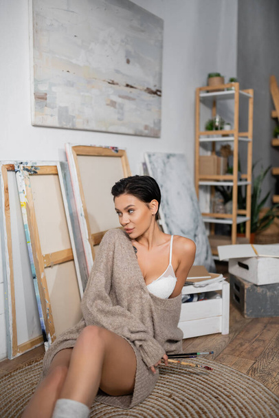 Sexy brunette artist in bra and sweater holding paintbrushes near blurred paintings in workshop  - Foto, Imagen