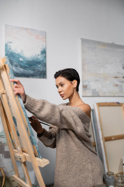 Sexy artist in sweater painting on canvas in workshop  - Foto, Imagen