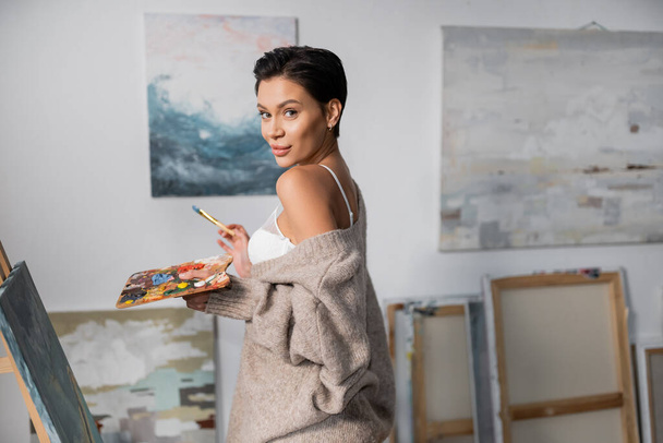 Brunette artist in bra and sweater holding palette with paints near canvas in workshop  - Fotografie, Obrázek
