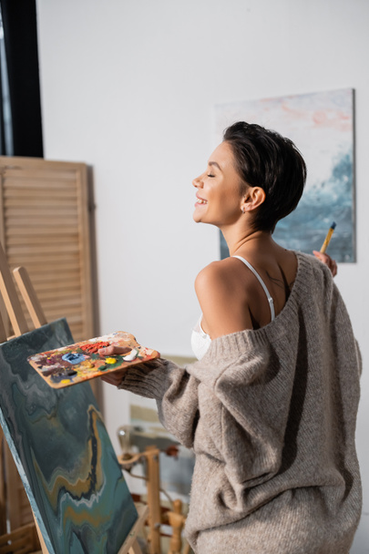 Πλευρική άποψη του χαρούμενου καλλιτέχνη στο σουτιέν και πουλόβερ κρατώντας παλέτα κοντά στη ζωγραφική στο εργαστήριο  - Φωτογραφία, εικόνα