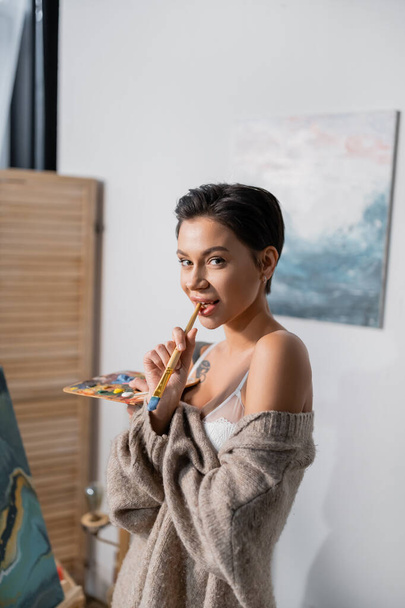 Sensual artist holding paintbrush near lips and looking at camera in studio  - Foto, Bild