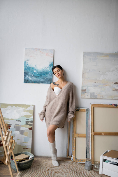 Seductive artist in sweater standing near paintings and easel in workshop  - Foto, Imagen