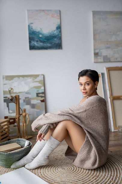 Short haired artist in socks and sweater looking at camera while sitting on floor in workshop  - Foto, Imagen