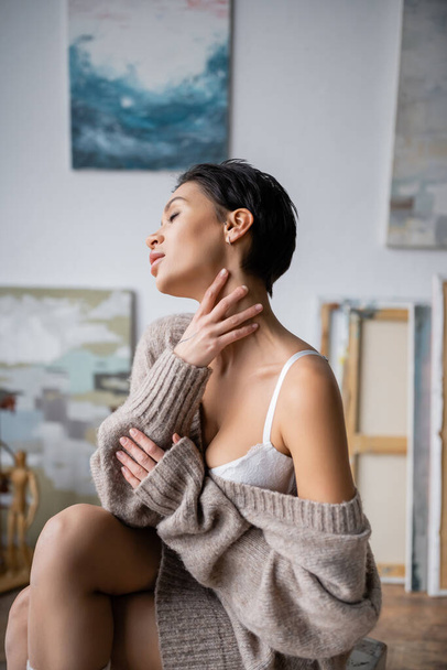 Seductive artist in sweater and bra touching neck in workshop  - Foto, imagen