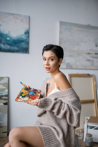 Αισθησιακός καλλιτέχνης σε πουλόβερ κρατώντας παλέτα και πινέλο στο εργαστήριο  - Φωτογραφία, εικόνα