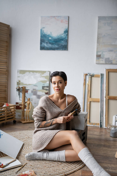 Tattooed artist in socks and sweater holding paintbrush while sitting on floor in workshop  - Valokuva, kuva