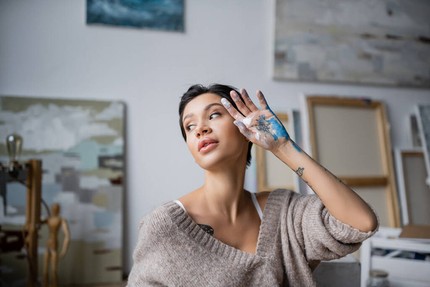 Σέξι καλλιτέχνης με το χέρι στο χρώμα κοιτάζοντας μακριά στο εργαστήριο  - Φωτογραφία, εικόνα