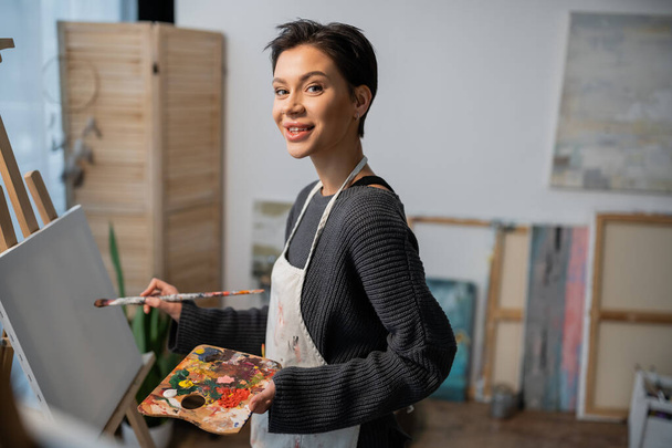 Smiling artist looking at camera while painting on canvas in studio  - Fotó, kép