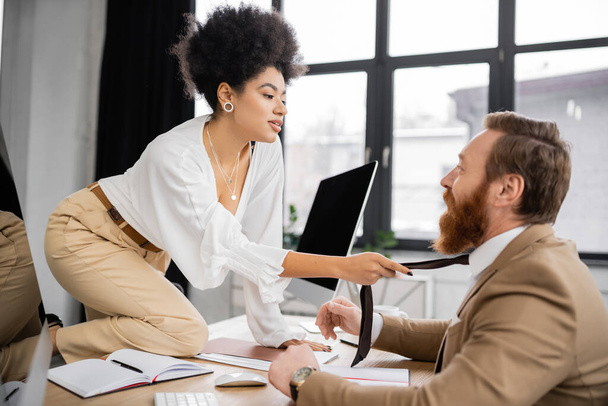 african american businesswoman pulling tie while seducing bearded coworker in office  - Foto, imagen