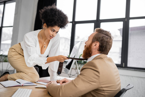 seductive african american woman sitting on desk and pulling tie of bearded coworker in office  - Foto, Imagem