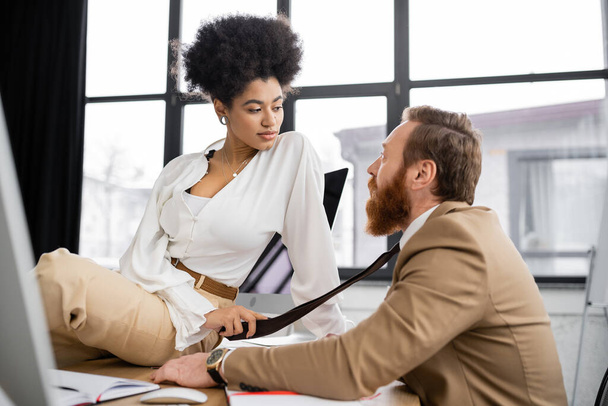 african american woman sitting on desk and pulling tie of coworker in office  - Foto, immagini