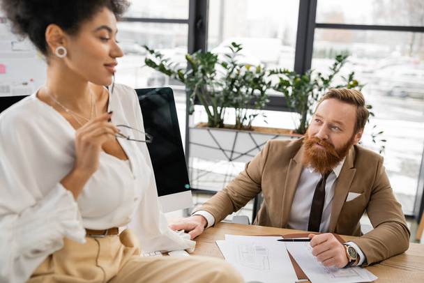cheerful and bearded man looking at seductive african american businesswoman holding glasses while sitting on desk  - Photo, Image