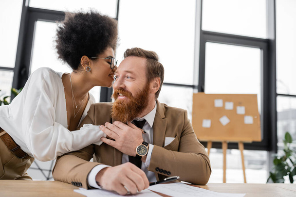 smiling african american businesswoman hugging while whispering in ear of cheerful coworker in office  - Foto, Bild