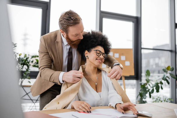 bearded businessman wearing jacket on pleased african american woman in office  - Photo, Image