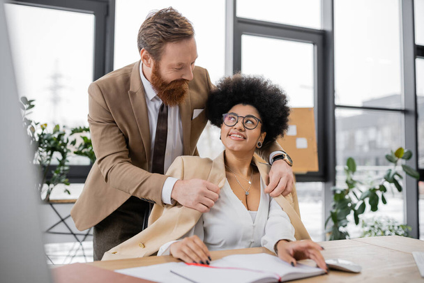 bearded businessman wearing jacket on cheerful african american woman in office   - Photo, image