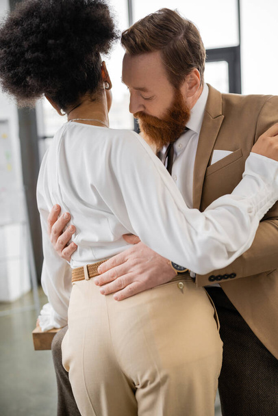 bearded man hugging curly african american colleague in office  - Foto, afbeelding