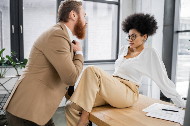 african american woman sitting on office desk and seducing bearded man pulling tie - Zdjęcie, obraz