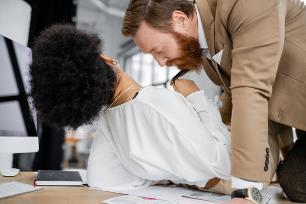curly african american woman pulling tie of bearded coworker while lying on office table - Fotoğraf, Görsel