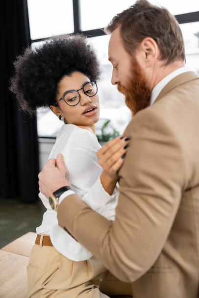 tattooed man touching shirt while undressing curly african american woman in office  - Photo, Image