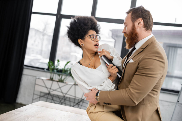 bearded businessman in formal wear trying to seduce scared african american colleague with notebook  - Photo, Image