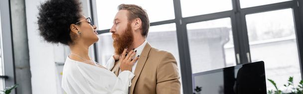 curly african american woman seducing bearded businessman near computer monitor in office, banner  - Foto, afbeelding