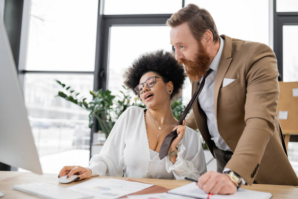 cheerful african american woman pulling tie of bearded businessman in office  - Photo, Image