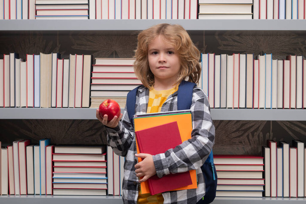 Koulupoika, jolla on kirjoja ja omena kirjastossa. Lapsi ala-asteelta kirjan kanssa. Nörttioppilas, joka on valmis opiskelemaan. Ensimmäinen kerta koulussa. Koulutuksen ja oppimisen käsite - Valokuva, kuva