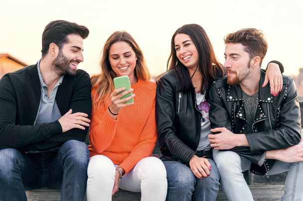 Happy group of trendy young people laughing and chatting sitting in a bench at sunset using a cellphone. Millennial friends having fun together outdoor - people, social network lifestyle concept - Foto, imagen