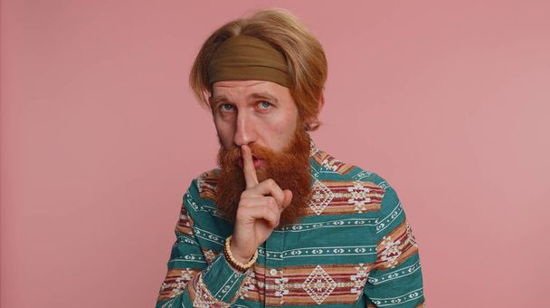 Shh be quiet please. Portrait of bearded redhead hippie man 20 years presses index finger to lips makes silence gesture sign do not tells secret. Young handsome hipster guy on pink studio background - Photo, Image