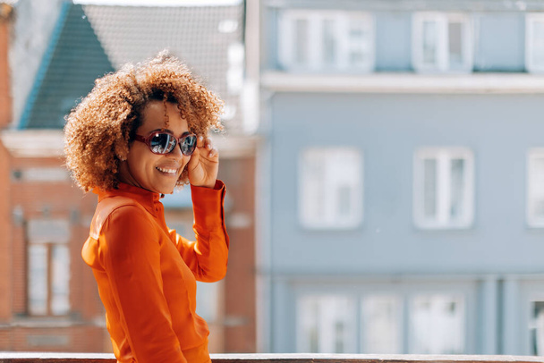 portrait beautiful smiling curly hair Brazilian young lady wearing sunglasses and orange color shirt looking in camera - outside home terrace picture in sunny daylight - Foto, imagen