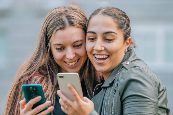 друзья с мобильными телефонами улыбаются обнимаясь на улице - Фото, изображение