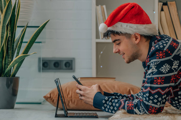 jovem com computador e telefone celular em casa no Natal com chapéu de santa claus - Foto, Imagem