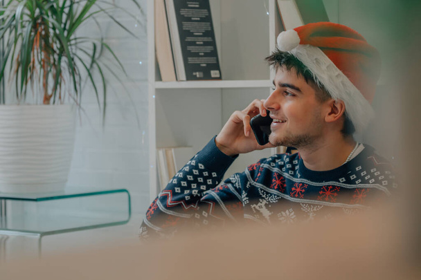 νεαρός άνδρας στο σπίτι με καπέλο Άγιος Βασίλης και χριστουγεννιάτικα ρούχα μιλώντας στο κινητό τηλέφωνο - Φωτογραφία, εικόνα