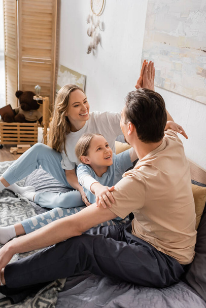 cheerful kid looking at father giving high five to happy mother while sitting on bed  - Valokuva, kuva