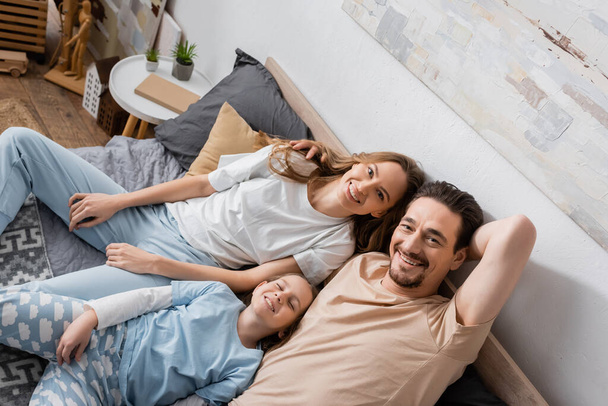 high angle view of happy parents lying with cheerful daughter on bed and looking at camera  - Foto, Imagen