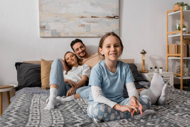 веселая девушка сидит на кровати рядом с размытыми родителями отдыхает на заднем плане  - Фото, изображение