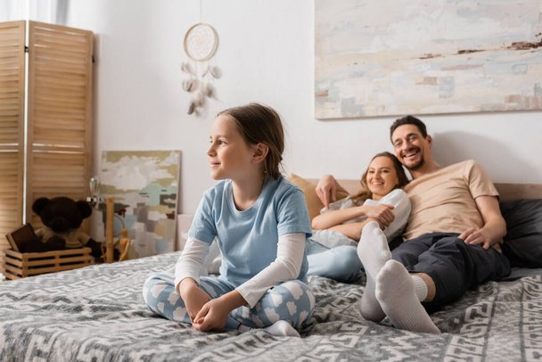 cheerful girl sitting on bed and looking away near blurred parents resting on background  - Foto, immagini