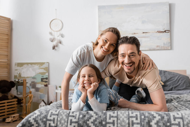 portrait of happy parents and kid looking at camera while resting in bedroom  - Fotoğraf, Görsel